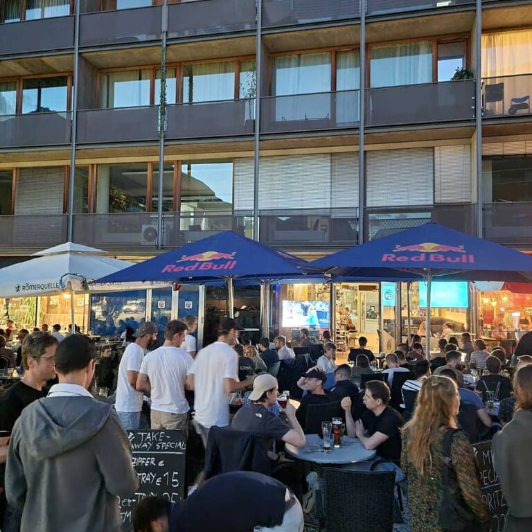 Group of friends socializing and enjoying drinks at the outdoor seating area of Café Bar Dinzler in Innsbruck, Austria, located near the SOWI campus, offering a cozy and vibrant atmosphere with wooden tables and ambient lighting Group of friends socializing and enjoying drinks at the outdoor seating area of Café Bar Dinzler in Innsbruck, Austria, located near the SOWI campus, offering a cozy and vibrant atmosphere with wooden tables and ambient lighting