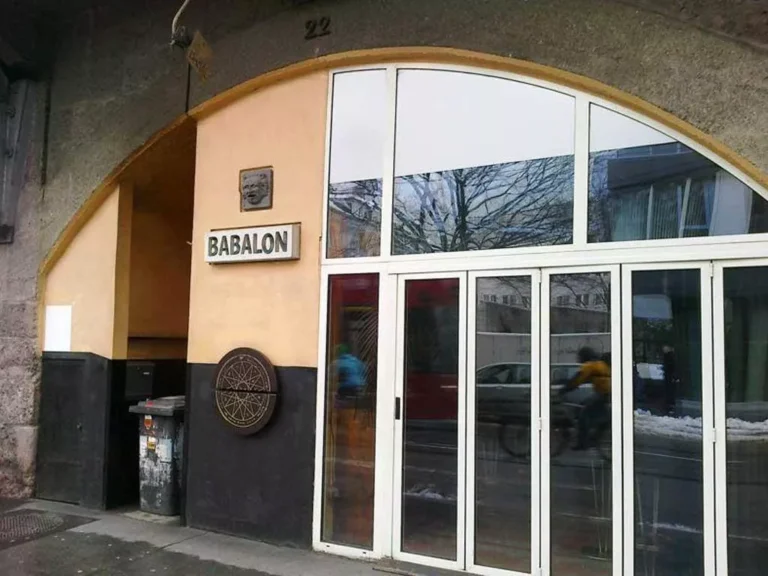 Entrance of Café-Bar Babalon in Innsbruck, Austria, located at Viaduktbögen 22, featuring a welcoming façade beneath the railway arches, with illuminated signage and a cozy outdoor seating area, offering a relaxed atmosphere for evening socializing