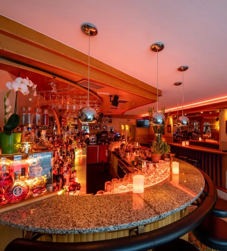 Bar area of M+M Bar in Innsbruck, Austria, featuring a sleek, illuminated counter with mosaic tiles, high stools, and a well-stocked selection of spirits, creating a stylish and inviting atmosphere for cocktail enthusiasts
