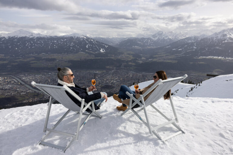 Guests relaxing with drinks on the snow-covered Cloud 9 Sun Terrace in Innsbruck, Austria, with the majestic Nordkette Alps providing a stunning backdrop under a clear blue sky